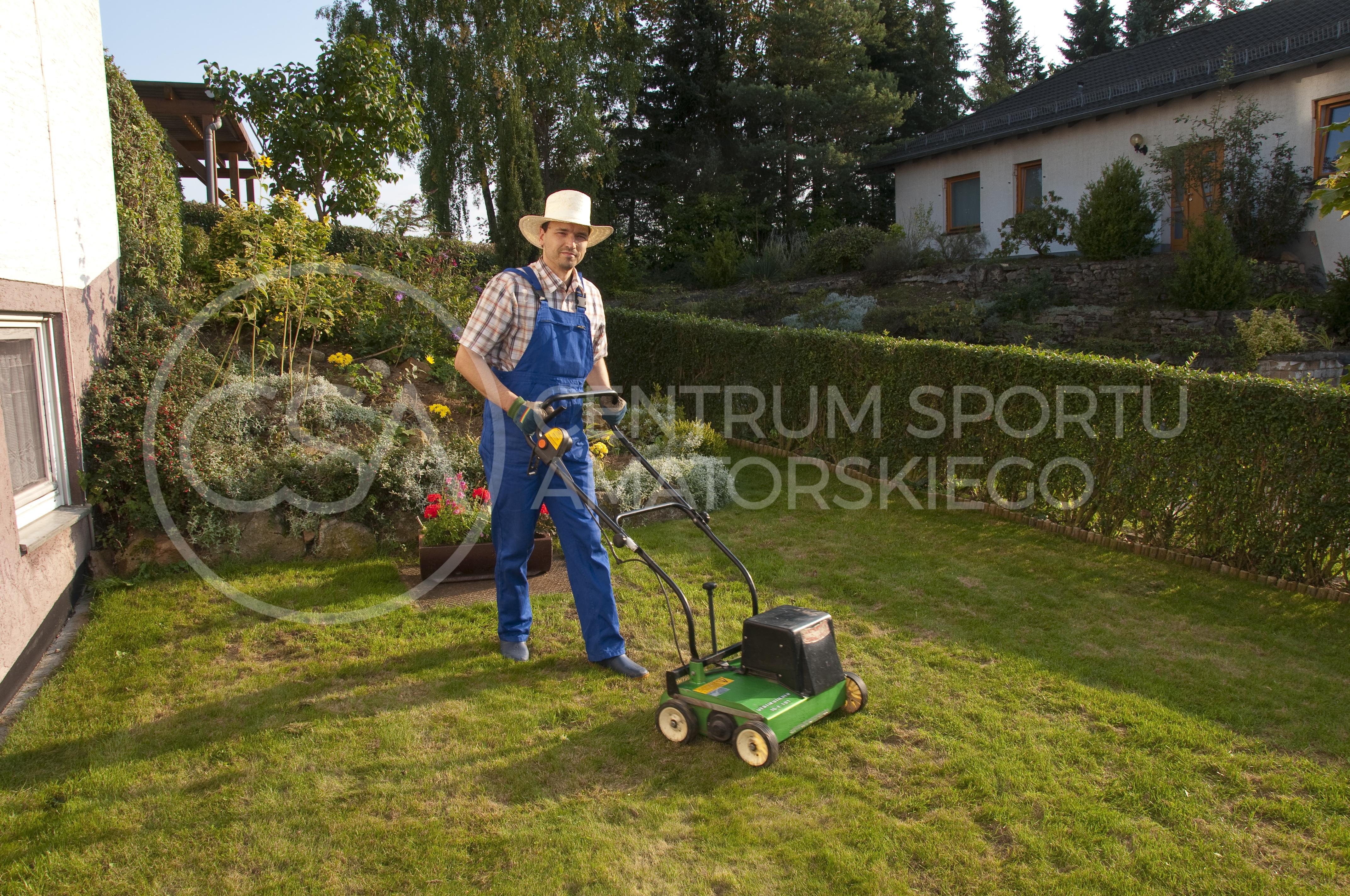 Man with blue overalls and a plaid shirt and straw hat standing in the garden and maintains the lawn with an electric scarifier.